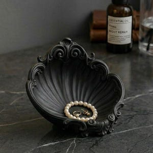 May include: A black, shell-shaped decorative dish with intricate detailing. A pearl bracelet and earrings are inside the dish. The dish sits on a dark, marble-effect surface, with other decorative items in the background.