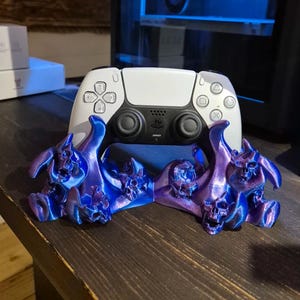Może przedstawiać: Biały kontroler PlayStation 5 umieszczony jest w stojaku w kształcie czaszki, wydrukowanym w technologii 3D. Stojak jest w żywym kolorze fioletowym i niebieskim.