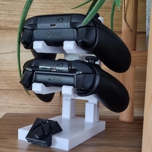 May include: A white plastic stand with two black Xbox controllers resting on it. The stand has a small Xbox logo on the front.