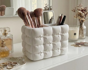 May include: A white, quilted makeup brush holder with bow accents, filled with makeup brushes. The vanity also features perfume bottles, a jewelry tray with rings and necklaces, and a small vase with dried flowers. A candle and mirror are also visible.