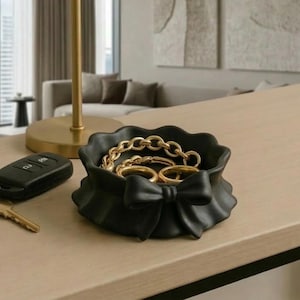 May include: A black decorative bowl with a bow design, filled with gold jewellery. A car key and remote are next to the bowl on a light-coloured wooden surface. The background includes a window and modern decor.