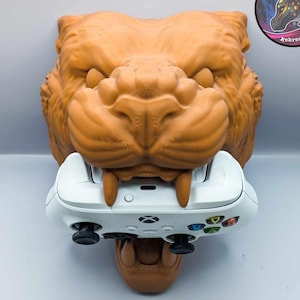 Może przedstawiać: Brązowa, trójwymiarowa głowa zwierzęcia z białą konsolą do gier wideo w pysku. Głowa zwierzęcia ma groźny wyraz twarzy z ostrymi zębami. Konsola ma przyciski i joystick.