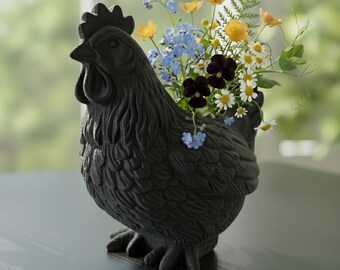 May include: A black, textured ceramic hen-shaped planter filled with a variety of colorful flowers. The flowers include yellow, blue, white, and purple blooms. The planter is on a dark surface.