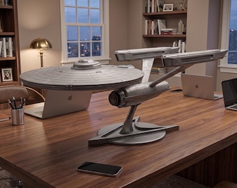 May include: A detailed gray model of a starship featuring a large circular saucer section, two elongated engine nacelles, and a central support stand, positioned on a wooden desk alongside two silver laptops and a black smartphone.