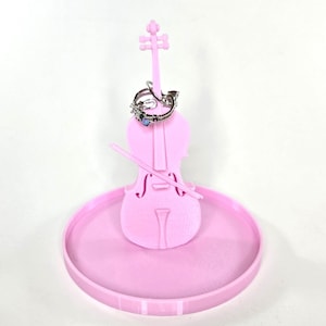 May include: A pink 3D printed cello ring holder with a ring on the top of the cello. The cello is sitting on a round pink base.