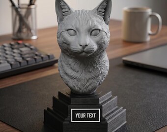 May include: A gray cat bust sculpture with detailed fur texture, mounted on a black stepped base. The base features a rectangular label with the text "YOUR TEXT" in a white font. The sculpture is on a desk.