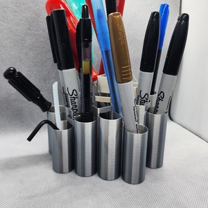 Puede incluir: Un portaplumas de metal plateado con múltiples ranuras, que contiene una variedad de bolígrafos, incluidos marcadores permanentes negros Sharpie, un marcador permanente azul Sharpie, un marcador permanente dorado Sharpie y un bolígrafo negro y rojo.