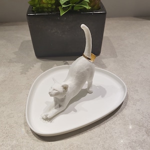 Pode incluir: Porta-anéis de cerâmica branca em forma de gato com uma faixa dourada em volta da cauda. O gato está em uma pose esticada, com a cabeça virada para o lado. O porta-anéis está em um prato de cerâmica branca.