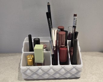 Puede incluir: Un organizador de maquillaje blanco con un patrón de diamante contiene varios artículos de maquillaje, incluidos lápices labiales, delineadores de labios y lápices de ojos. El organizador tiene varios compartimentos para guardar diferentes artículos.