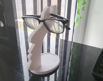 May include: White 3D printed face shaped eyeglasses holder with a pair of black framed glasses resting on it.