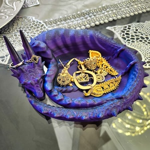Puede incluir: Un plato de dragón morado para baratijas con joyas de oro en su interior. El plato está hecho de un material impreso en 3D y tiene una superficie texturizada.