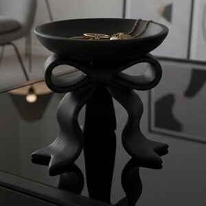 May include: A black decorative bowl with a bow-shaped base, holding jewellery, sits on a black table. The bowl is round and has a smooth surface. The background includes a grey chair, a lamp, and framed abstract art.