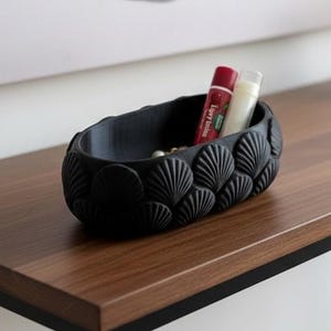 Könnte beinhalten: Eine schwarze, muschelförmige Dekoschale mit zwei Tuben auf einem Holztisch. Der Tisch hat ein schwarzes Metallgestell. Eine kleine Sukkulente in einem geometrischen Topf und eine Lampe befinden sich ebenfalls auf dem Tisch.