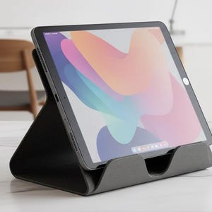 May include: A black tablet rests on a dark grey stand. The tablet screen displays a colourful abstract design. Two small succulents in white geometric pots are on the right. A grey desk lamp and wooden chair are in the background.