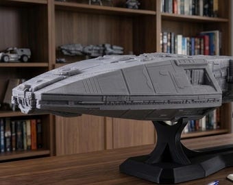 May include: A large, gray, detailed model of a spaceship resting on a black stand. The vessel features a long, angular hull with intricate paneling and a small figurine standing on its side. The model is displayed on a wooden surface.