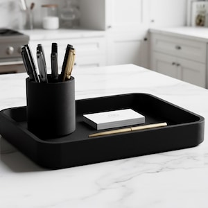 Pode incluir: Uma bandeja retangular preta com cantos arredondados contém um porta-canetas preto com canetas, uma pilha de cartões de visita brancos e uma caneta dourada. Os itens estão em uma bancada de mármore branco.