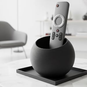 Puede incluir: Un mando a distancia gris se encuentra en un soporte esférico negro sobre una bandeja rectangular negra. El soporte está sobre una mesa de mármol blanco. El mando tiene varios botones y un control circular.