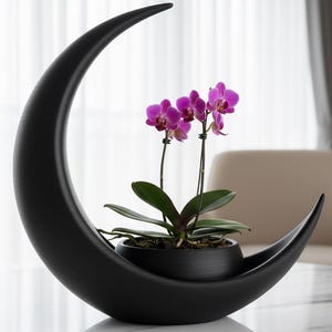 Könnte beinhalten: Ein schwarzer, halbmondförmiger Pflanztopf mit einer Topf-Orchidee mit leuchtend violetten Blüten. Der Pflanztopf hat eine glatte, matte Oberfläche und steht auf einer weißen Marmoroberfläche. Die Orchidee hat grüne Blätter.