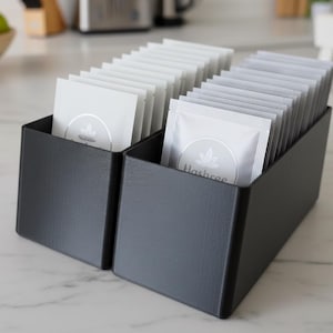 Op de afbeelding: Twee zwarte rechthoekige organizers gevuld met witte zakjes. Elk zakje heeft een zilveren logo en tekst. De organizers staan op een wit marmeren oppervlak. De achtergrond omvat een fruitschaal en keukenapparatuur.