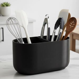 Puede incluir: Un soporte negro para utensilios de cocina con varios compartimentos, lleno de diversas herramientas de cocina. Incluye un batidor, espátulas, pinzas y una cuchara de madera. El soporte está sobre una encimera blanca.