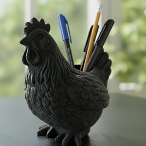 May include: Black rooster-shaped desk tidy holding pens and pencils. The organiser is made of a textured material and features the text "Black Color". The rooster has a detailed comb, wattle, and feather design. A unique and functional desk accessory.