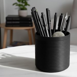 May include: A black cylindrical desk tidy with horizontal ridged detailing, holding various black pens and brushes. A small, grey stone sits inside the tidy. The tidy is on a white table, with a plant and books in the background.