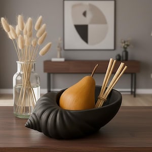 Peut inclure: Un bol décoratif noir en forme de coquillage contient une poire et un bouquet de bâtonnets en bois. Un vase en verre transparent avec des fleurs séchées est à gauche. La scène se déroule sur une table en bois foncé.