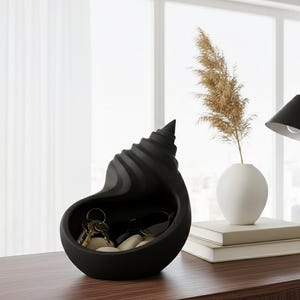 May include: A black, shell-shaped key holder containing keys and small stones. A white vase with dried plants, a black desk lamp, and two books sit on a wooden table. A neutral armchair is in the background.