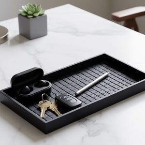 May include: A black rectangular tray with a textured surface, holding a pair of wireless earbuds in a case, keys, a car key fob, and a silver stylus. A small succulent in a gray square pot is in the background.