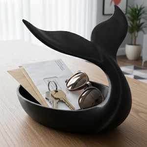 Puede incluir: Una bandeja decorativa negra con forma de cola de ballena que contiene llaves, gafas de sol y sobres. La bandeja está sobre una mesa de madera. La cola se curva hacia arriba, creando un almacenamiento único.