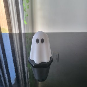 Puede incluir: Una figurita de fantasma blanca con ojos negros, sentada sobre una superficie negra.
