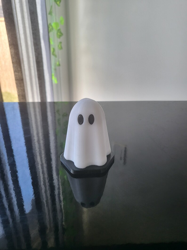 Puede incluir: Una figurita de fantasma blanca con ojos negros, sentada sobre una superficie negra.