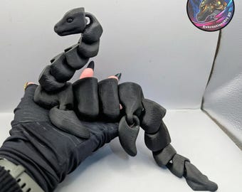 May include: A black articulated scorpion hand accessory. The scorpion is made of segmented pieces, allowing for movement. The accessory is worn on the hand, with the scorpion's tail extending beyond the wrist. The product is 3D printed.