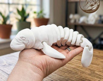 May include: A white, articulated manatee figurine with a segmented body design that allows for flexible movement. The creature features a rounded head, small eyes, and flat flippers, showcasing a smooth, matte finish throughout its entire structure.