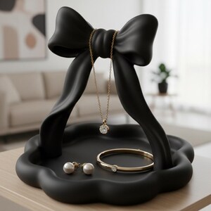 以下が含まれることがあります： 黒いリボン型のジュエリーオーガナイザー。ゴールドのペンダントネックレス、ゴールドのブレスレット、パールイヤリングを収納。明るい色の木製テーブルの上に置かれています。