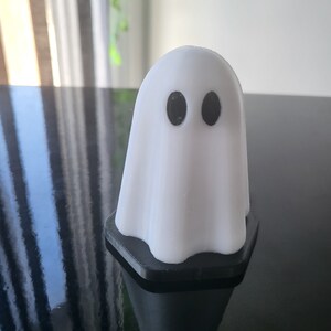 Puede incluir: Una figurita de fantasma blanca con ojos negros, de pie sobre una base hexagonal negra.