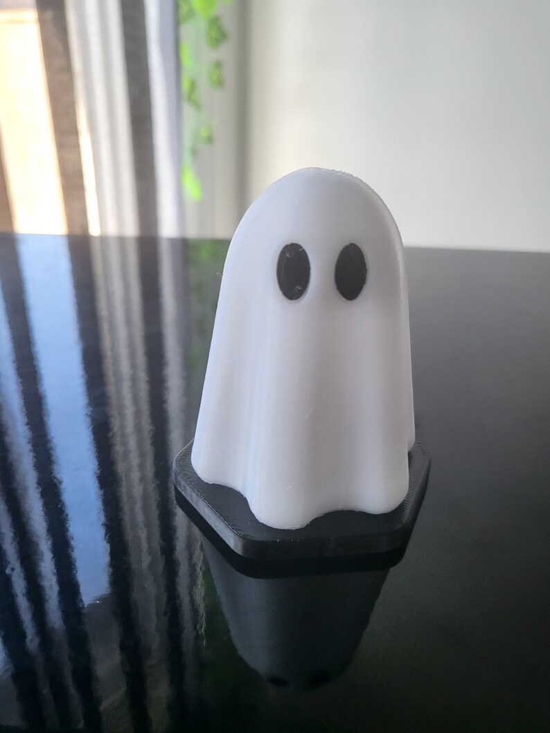 Puede incluir: Una figurita de fantasma blanca con ojos negros, de pie sobre una base hexagonal negra.