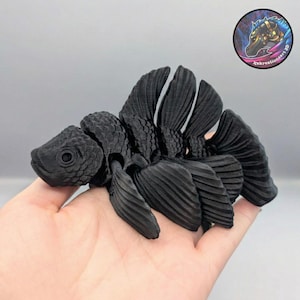 Peut inclure: Une sculpture de poisson articulée noire avec des nageoires et des segments de corps détaillés. La sculpture est tenue dans une main, mettant en valeur son design complexe. Un logo circulaire avec une tête de cheval est visible en arrière-plan.