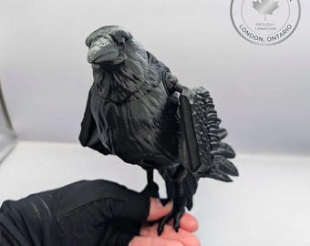 May include: A matte black articulated raven figurine featuring textured feather details on the wings and body. The bird has a sharp beak and visible joints at the wing attachments. A circular logo in the corner reads Made in Canada London Ontario.