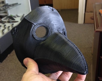 May include: A black plastic plague doctor mask with a long beak and a round eye hole. The mask is textured and has a stitched seam around the edge.