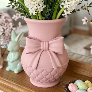 May include: A pink ceramic vase with a bow detail, filled with white flowers. Two ceramic bunny figurines and a basket of decorative eggs are also visible, suggesting an Easter theme.