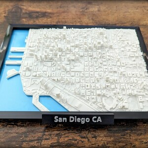 May include: A 3D printed model of the San Diego skyline in a black frame with a blue base. The model is made of white plastic and shows the city's buildings and streets. The frame has a label that reads "San Diego CA".