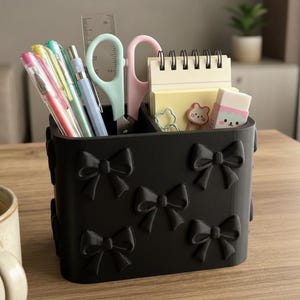 May include: A black desk organiser with bow accents, filled with pens, scissors, a ruler, and a notepad. The organiser is on a wooden surface, with a coffee mug in the foreground. The organiser is approximately 10 cm wide.