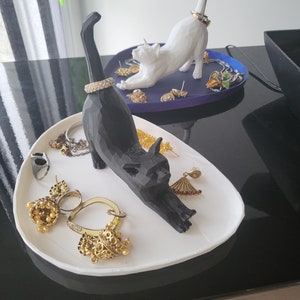 May include: Two 3D printed cat ring holders, one black and one white, on white and blue trays. The trays are filled with jewelry, including rings, earrings, and necklaces.