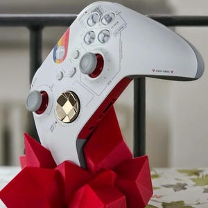 Op de afbeelding: Een witte en rode Xbox-controller met een gouden accent op de directionele pad wordt weergegeven op een rode geometrische standaard. De controller heeft een regenboogontwerp aan de voorkant en de tekst "GRIP HERE" aan de achterkant.