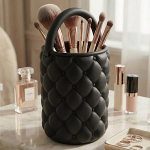 May include: A black, quilted makeup brush holder with a handle, filled with various makeup brushes. The holder is cylindrical and sits on a marble surface, alongside perfume and makeup products. The brushes have light-coloured handles and dark bristles.