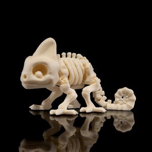 May include: A cream-coloured chameleon skeleton figurine. The figurine is posed in a walking position, with its rib cage and spine clearly visible. The tail is curled. The figurine is set against a black background, with a reflection.