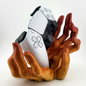 Pode incluir: Um controle branco e preto do PlayStation 5 repousa em um suporte em forma de mão laranja e marrom impresso em 3D. Os dedos do suporte embalam o controle, criando uma peça de exibição única para jogadores.
