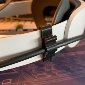May include: A black plastic cable clip attached to a white headband. The clip is designed to hold a cable in place.