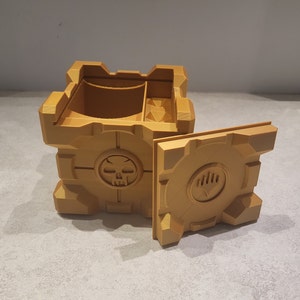 May include: A gold-colored 3D printed box with a skull and crossbones design on one side and a hand print on the other side. The box is open and has a compartment inside.
